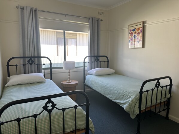 A room with two single beds, a small bedside table with a lamp, a window with curtains, and a framed picture on the wall. - O'Sea - Neat and tidy (Robe)