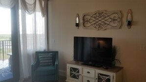 Een woonkamer met een grote flatscreen televisie op een houten kast, een decoratief kunstwerk erboven en een groene fauteuil.