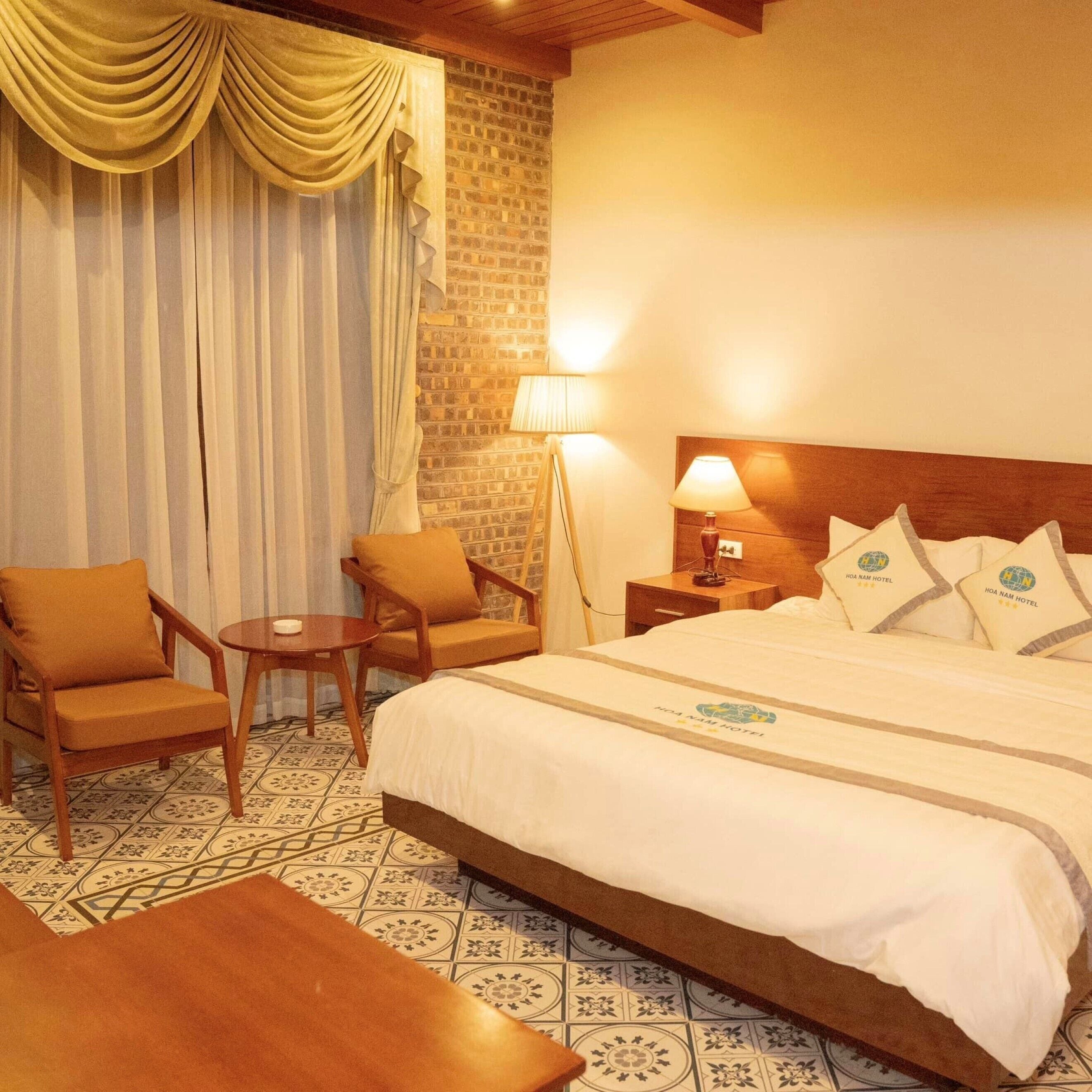 A hotel room with a bed, two armchairs, a small table, a nightstand with a lamp, and a brick wall.