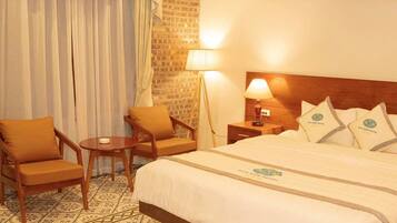 A hotel room with a bed, two armchairs, a small table, a nightstand with a lamp, and a brick wall.