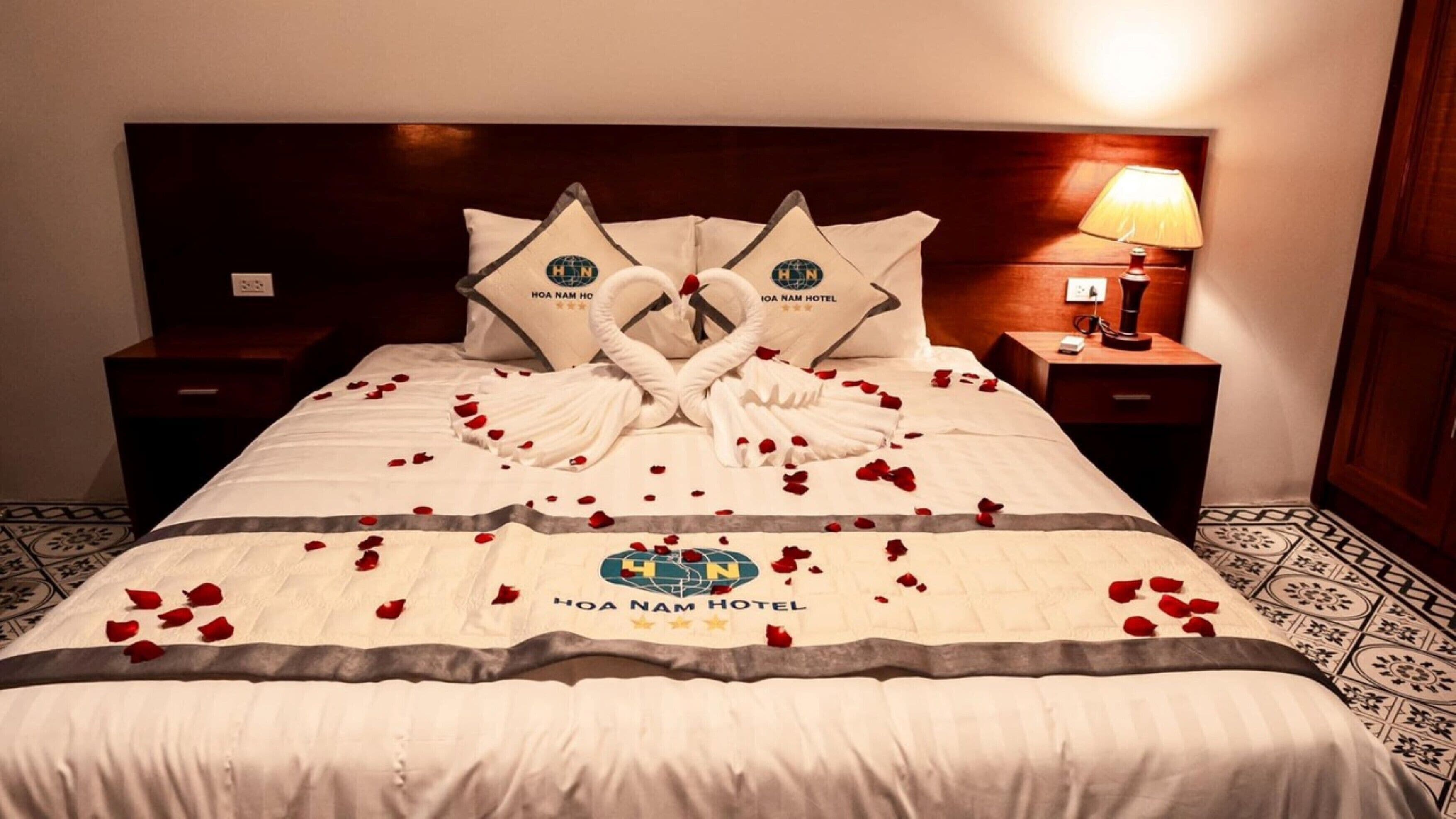 A hotel bed with a white bedspread, a logo in the center, and a towel arrangement forming a heart shape.