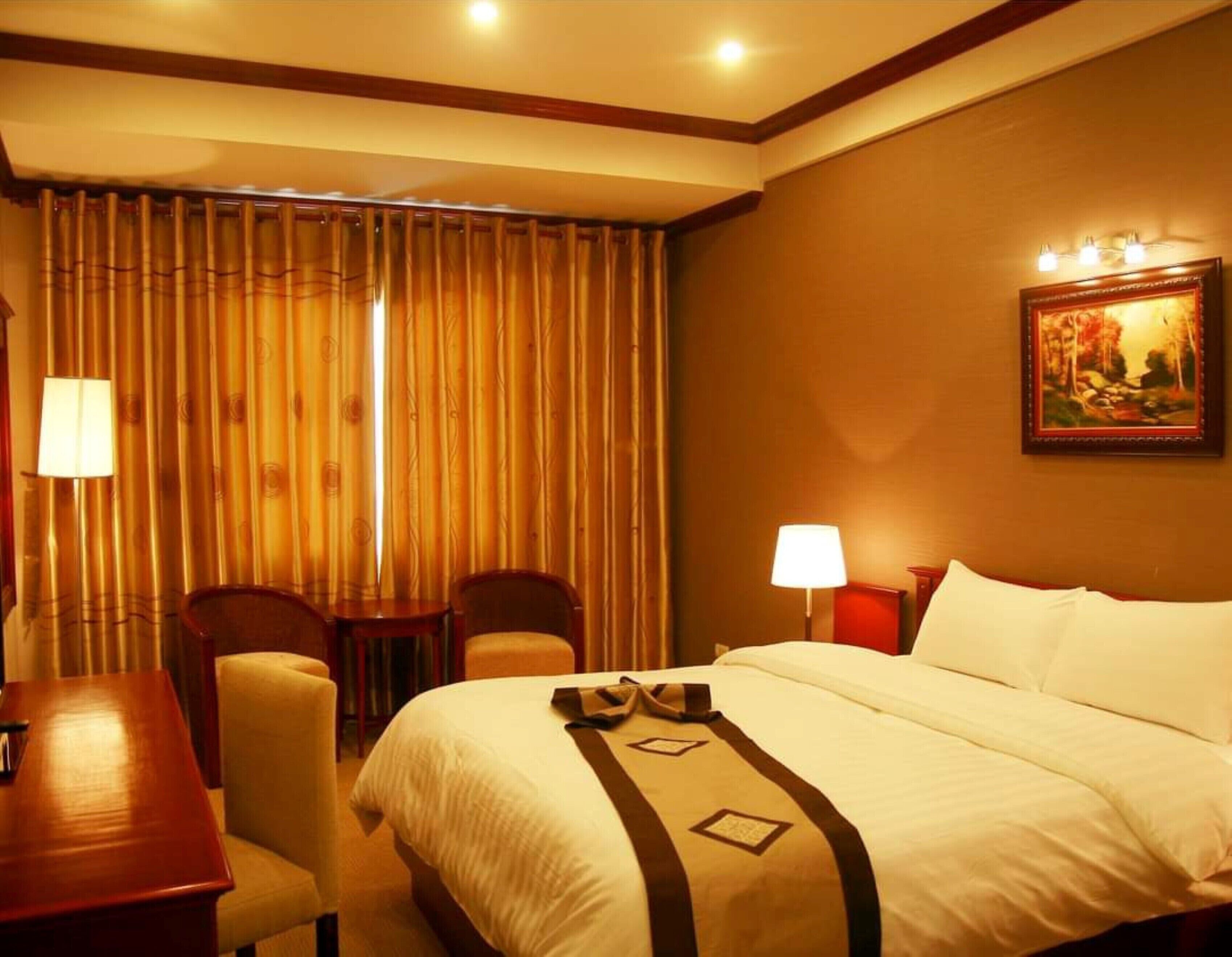 A hotel room with a bed, two chairs, a desk, and a painting on the wall.