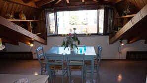 A dining area with a table and chairs, a window with a view of buildings, wooden beams, and shelves with items. - Beautiful Attic Apartment Near the Centre of Lignano Sabbiadoro (Lignano Sabbiadoro)