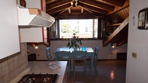 A kitchen with a gas stove, a dining table with chairs, a window with a view of buildings, and a wooden ceiling. - Beautiful Attic Apartment Near the Centre of Lignano Sabbiadoro (Lignano Sabbiadoro)