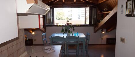 A kitchen with a gas stove, a dining table with chairs, a window with a view of buildings, and a wooden ceiling.