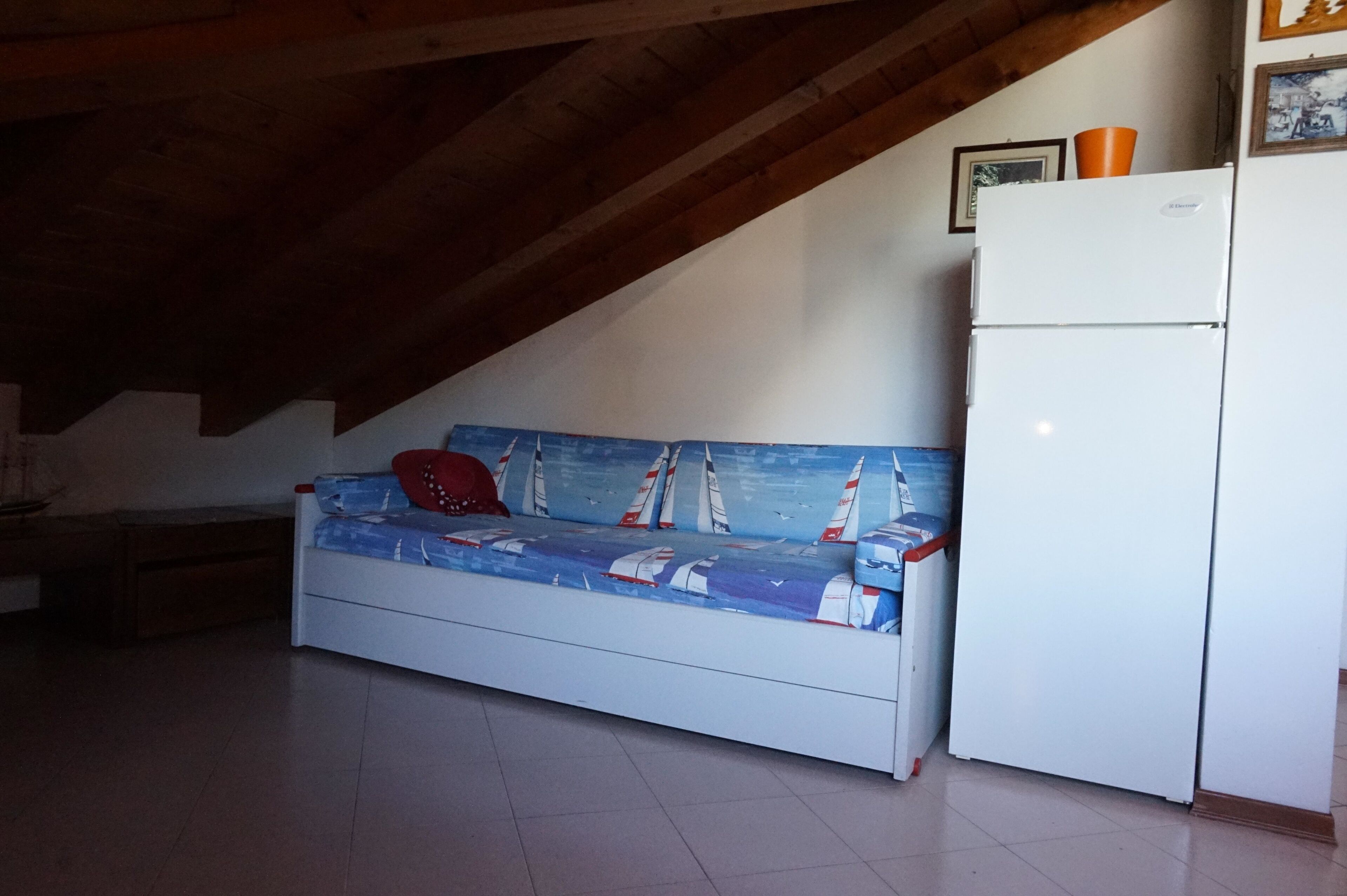 A room with a white sofa bed with a sailboat design, a white refrigerator, and wooden beams on the ceiling.