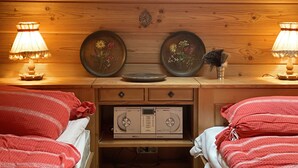 Une pièce chaleureuse aux murs en bois, avec deux lits dotés de draps rouges, deux lampes fixées au mur, une petite table sur laquelle est posée une assiette décorative, et un lecteur radio numérique.