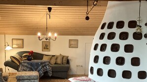 Un salon chaleureux avec un plafond en bois, un canapé vert agrémenté de coussins à motifs, une petite table sur laquelle est posé un vase, et un mur original orné d’ouvertures circulaires.