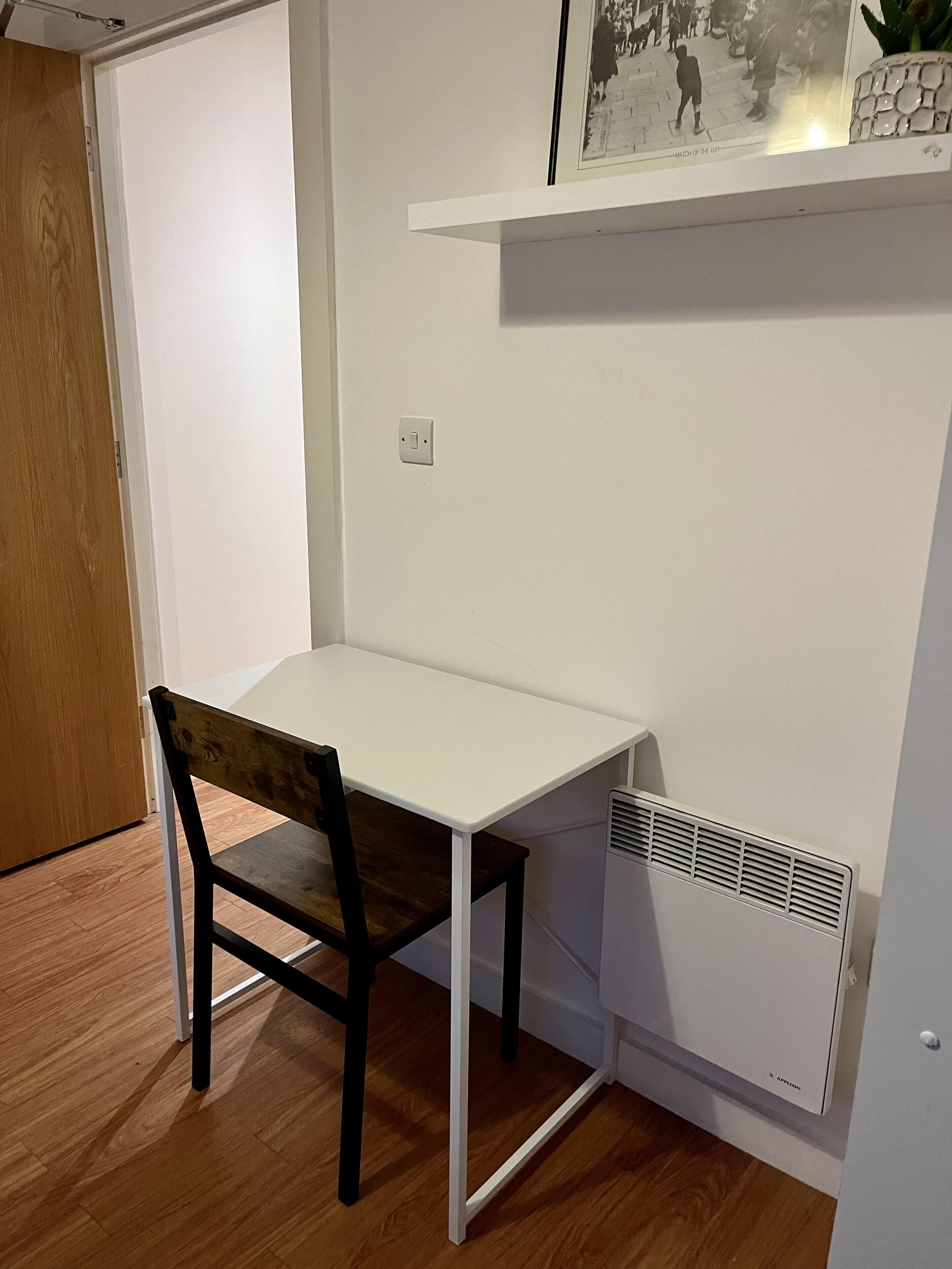 Uma pequena mesa branca com uma cadeira de madeira num quarto com soalho de madeira e parede branca.