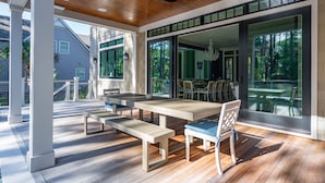 An outdoor patio with wooden tables and benches, a ceiling fan, and large sliding glass doors leading to a dining area. - Stunning Coastal Gem in Vanderhorst with private pool (Kiawah Island)