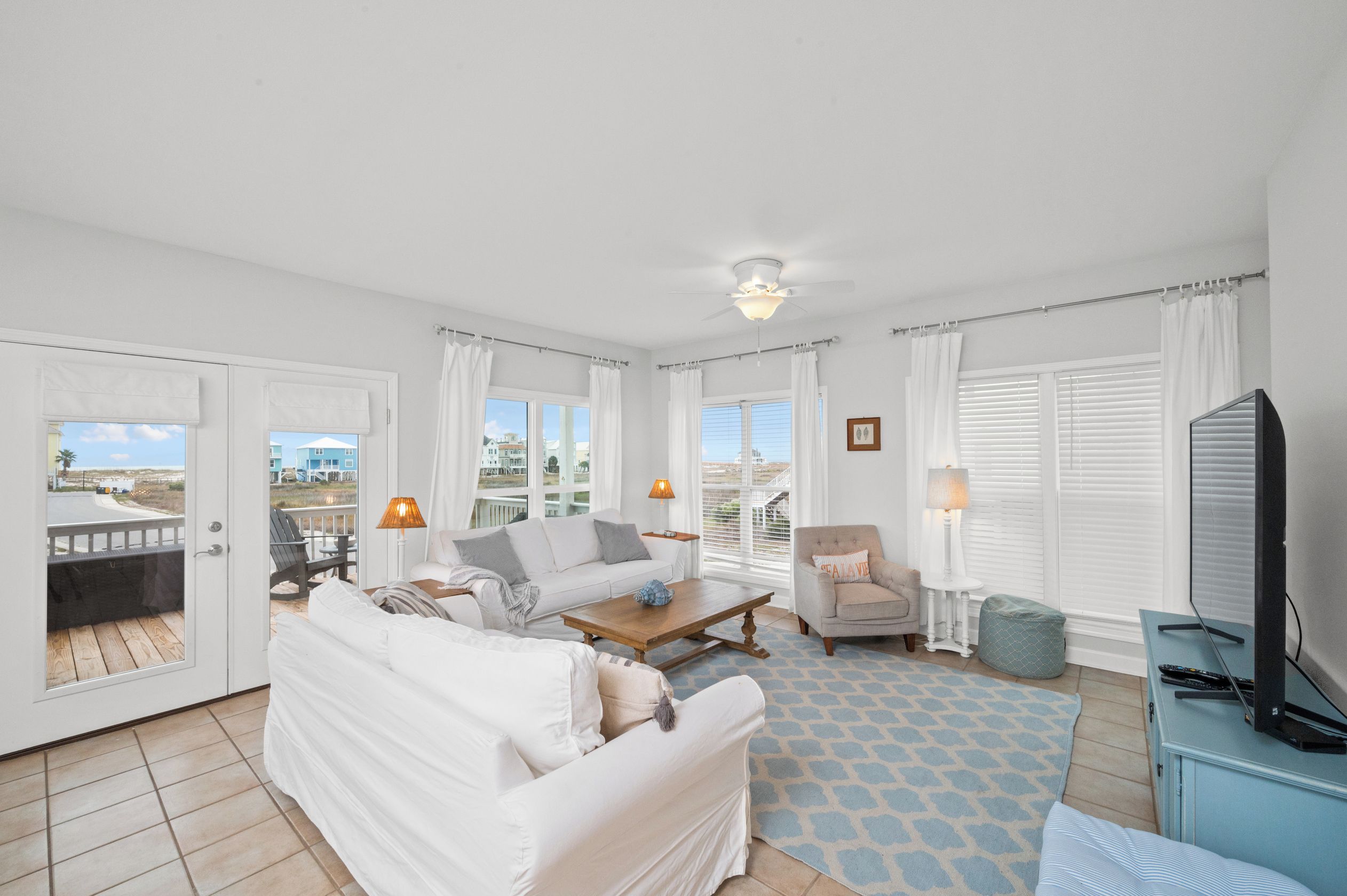 A spacious living room with a white sofa, a wooden coffee table, and a television on a blue cabinet. There are two windows with white curtains, and a balcony is visible through one of the windows.