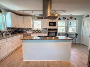 A modern kitchen with white cabinets, a central island, stainless steel appliances, and a large window providing natural light.