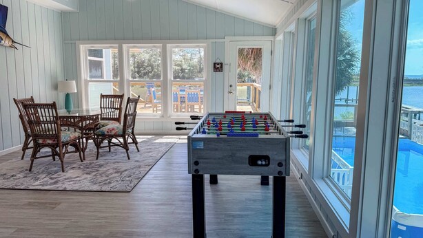 A room with a foosball table, wicker chairs, a glass table, and large windows overlooking a deck. - St.George Island Bayfront Paradise with Pool,Dock (St. Geroge Island)
