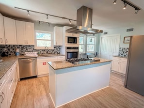 A modern kitchen with white cabinets, a stainless steel refrigerator, a built-in stove, and a large island with a granite countertop. - St.George Island Bayfront Paradise with Pool,Dock Golf Cart Included (St. Geroge Island)