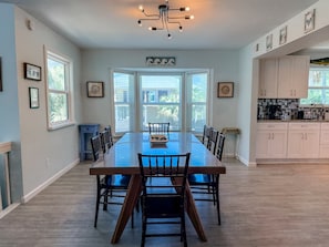 A spacious dining area with a long table and chairs, large windows, and a kitchen with white cabinets. - St.George Island Bayfront Paradise with Pool,Dock Golf Cart Included (St. Geroge Island)
