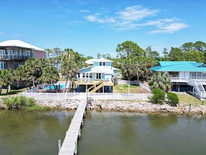 A coastal property with a dock leading to a house, surrounded by palm trees and a clear blue sky. - St.George Island Bayfront Paradise with Pool,Dock Golf Cart Included (St. Geroge Island)