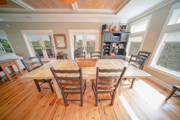 A dining area with a wooden table and chairs, a kitchen with cabinets and a countertop, and large windows with a view of greenery.