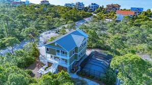 A two-story house with a blue roof, surrounded by dense greenery and a clear view of the sea. - Private Screened Pool, Easy Beach Access in Plantation! (St. George Island)