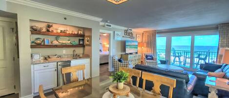 Amplia sala de estar con un ventanal con vista al océano, una mesa de comedor con sillas y una cocina con estantes decorativos.