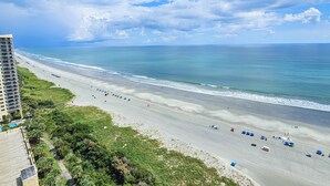A beach with a hotel in the background, a clear sky, and the ocean. - 1703 Margate Gorgeous oceanfront condo with full resort access! (Myrtle Beach)