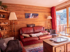 A cozy living room with a red leather sofa, wooden walls, and a large window with a view of trees.