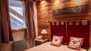 Camera da letto con testiera in legno, letto con copriletto a quadretti rossi e bianchi, due cuscini con motivi a cuore, comodino con lampada e finestra con tende a quadretti.