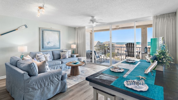 A spacious living room with a large window overlooking the beach, a dining area with a view, and a ceiling fan.