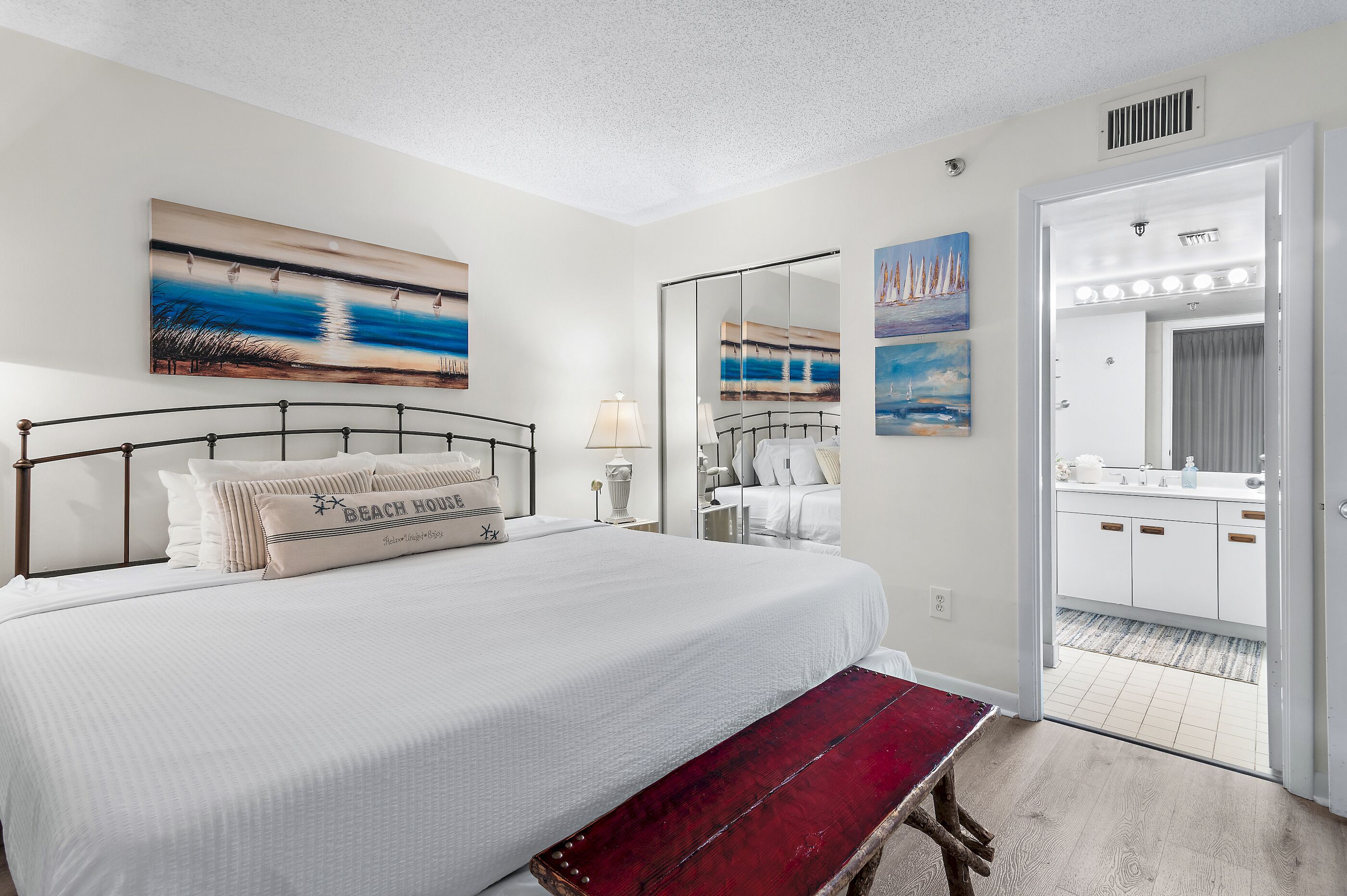 A hotel room with a large bed, a bench, a vanity with lights, and a door leading to a bathroom.