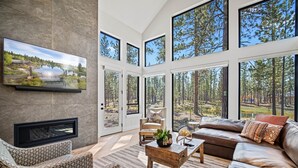 Ein großzügiges Wohnzimmer mit großem Fernseher, bequemen Sitzgelegenheiten und Blick auf einen Wald durch große Fenster.