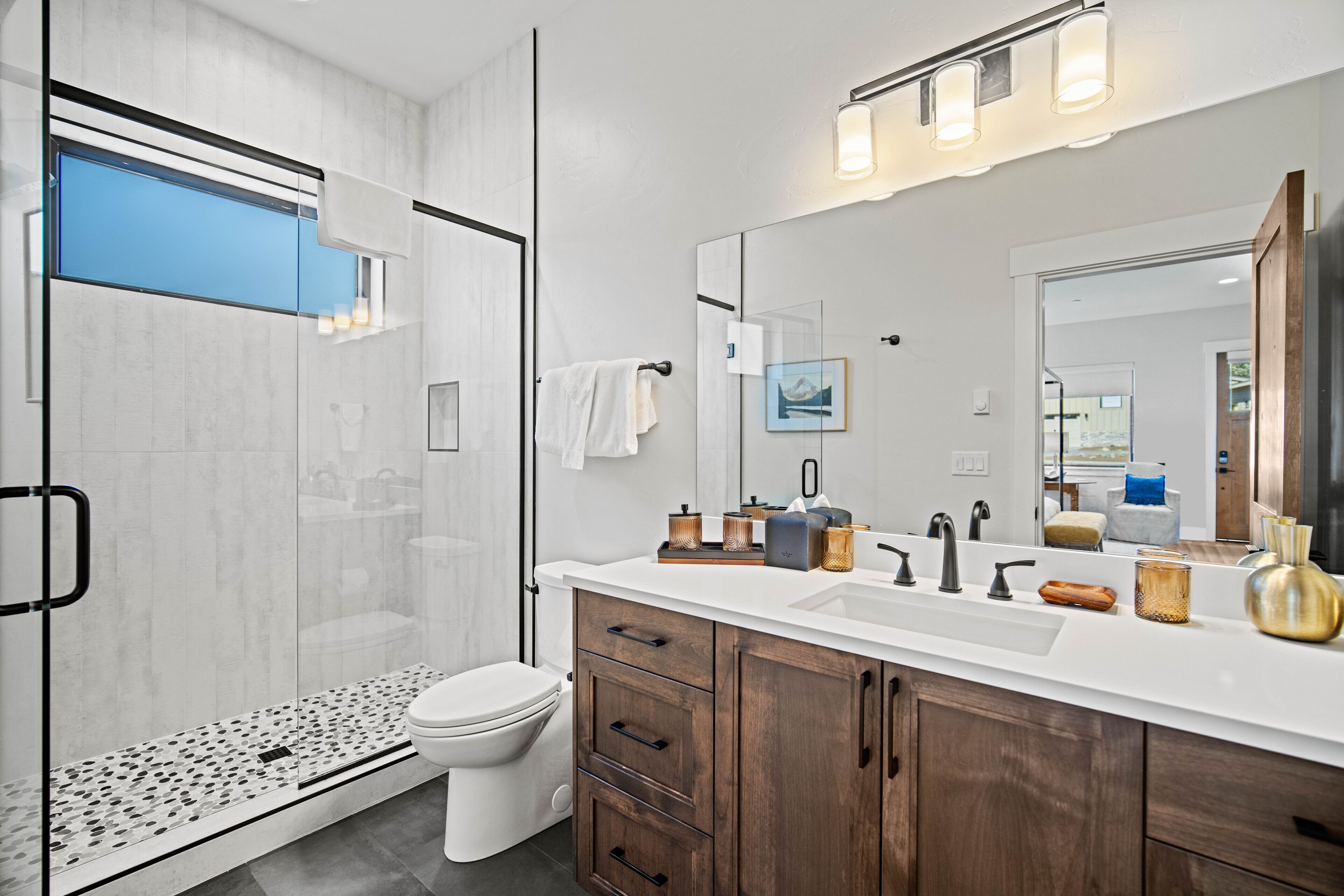 A modern bathroom with a glass shower enclosure, a white toilet, a large mirror with lights, and wooden cabinets.