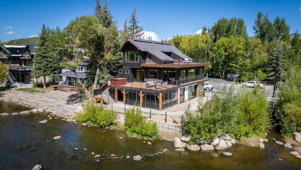 Una casa moderna con un balcón que da a un río, rodeada de árboles y un estacionamiento.