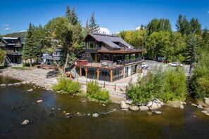 A modern house with a deck overlooking a river, surrounded by trees and a parking area.