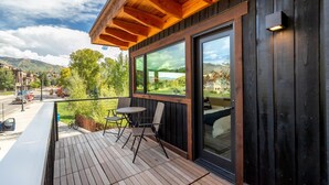Terraza de madera con mobiliario exterior, un ventanal grande y vistas a un paisaje montañoso.