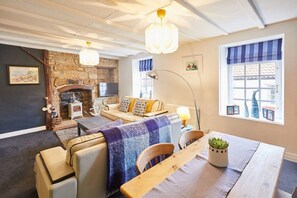 A cozy living room with a stone fireplace, a sofa, a dining table with chairs, and a large window with blue curtains. - Host & Stay | Dotty's Coastal Retreat (Staithes)