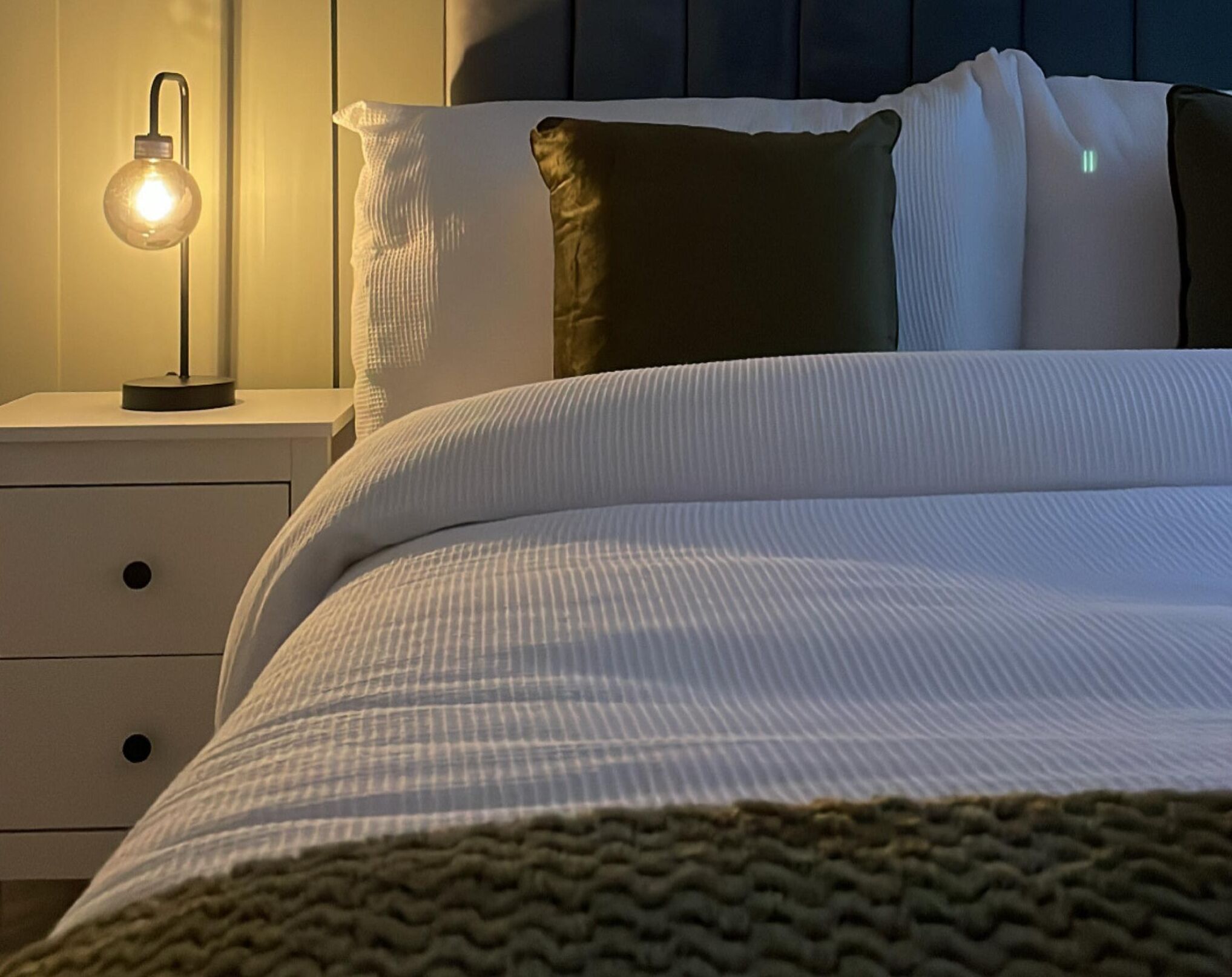 Ein ordentlich bezogenes Bett mit weißer und grüner Decke, einer Nachttischlampe und einem Kopfkissen auf einem weißen Nachttisch.