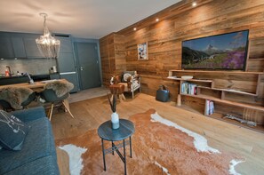 A modern living room with a wooden wall, a mounted TV, and a dining area with a chandelier. - Z Suite - Haus Roger (Zermatt)