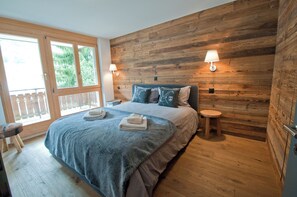 A bedroom with a wooden wall, a bed with a blue blanket, two bedside lamps, and a wooden stool. - Z Suite - Haus Roger (Zermatt)