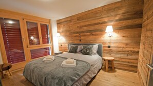 Ett sovrum med en väggpanel av trä, en säng med ett grått överkast, två sänglampor och en liten träpall.