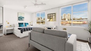 Une chambre moderne avec un lit, un canapé gris, un bureau blanc et de grandes fenêtres donnant sur la ville.