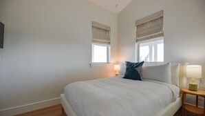 Een slaapkamer met een groot bed, twee nachtkastjes met lampen, en ramen met jaloezieën.