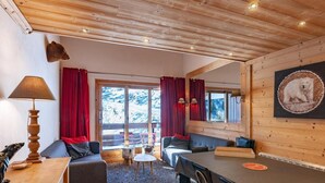 A cozy cabin interior with a wooden ceiling, a sofa, a dining table, and a large window with red curtains.
