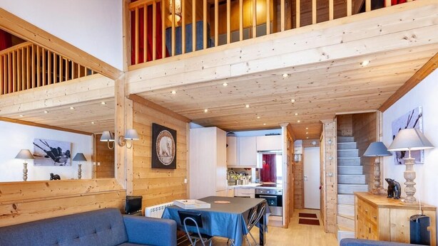 A spacious living area with a high ceiling, wooden beams, a dining table with chairs, and a kitchen in the background.