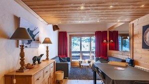 A cozy cabin interior with wooden walls and ceilings, a dining area with a table and chairs, a sofa, and a view of snowy mountains through large windows.