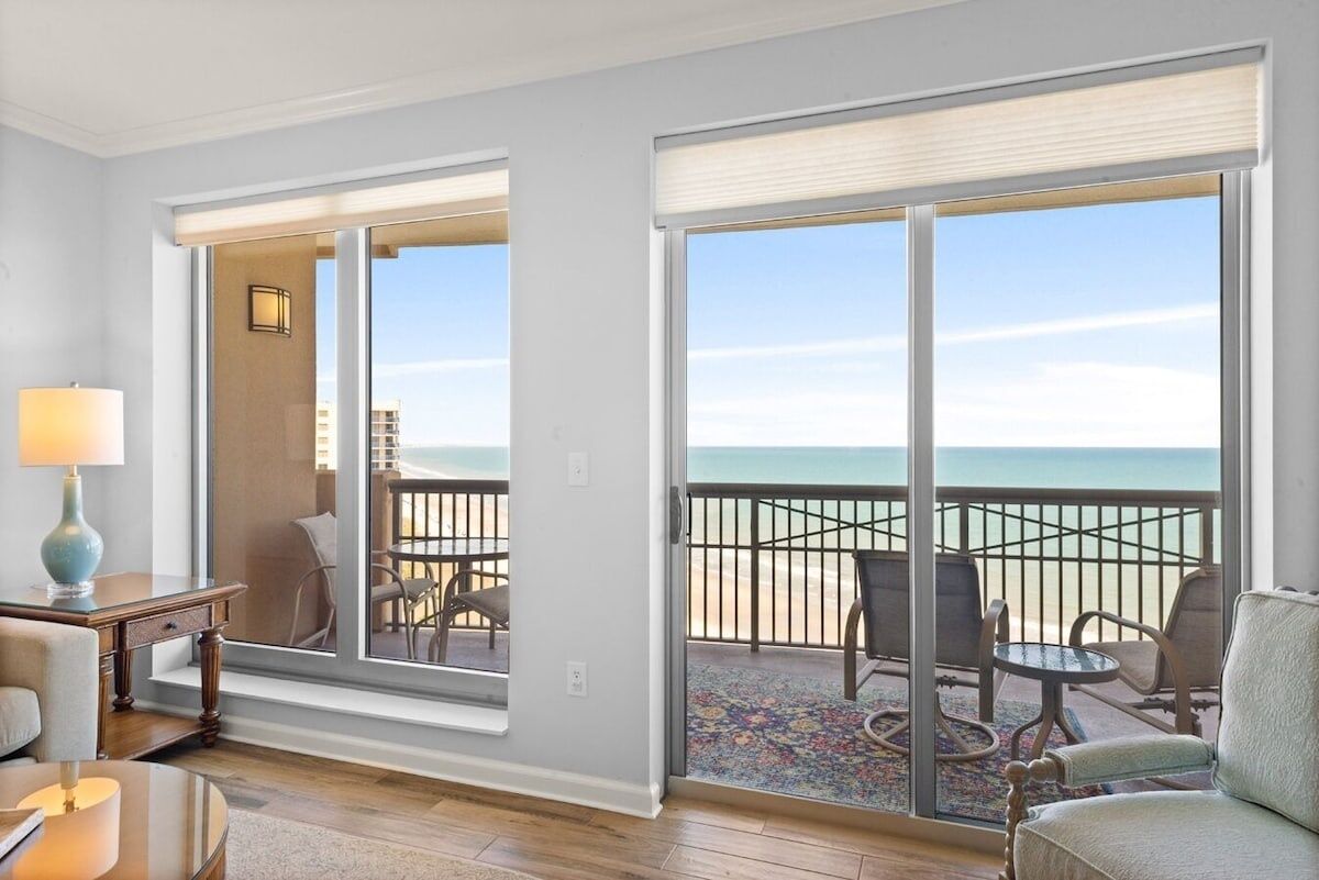 A room with a large sliding glass door leading to a balcony with a view of the ocean. There is a small table with a lamp and a chair inside the room.