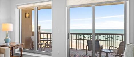 A room with a large sliding glass door leading to a balcony with a view of the ocean. There is a small table with a lamp and a chair inside the room.