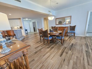 Un vaste espace de salle à manger avec une table en bois et des chaises en osier, une cuisine dotée de meubles blancs et un couloir menant aux autres pièces.