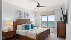 Ein Schlafzimmer mit einem hölzernen Bett, zwei Nachttischen mit Lampen, einem Deckenventilator, einem großen Fenster mit Meerblick und zwei Wandbildern.