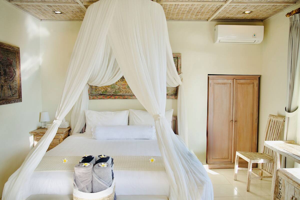 A bedroom with a four-poster bed, a wooden wardrobe, a bedside table with a lamp, and a chair.