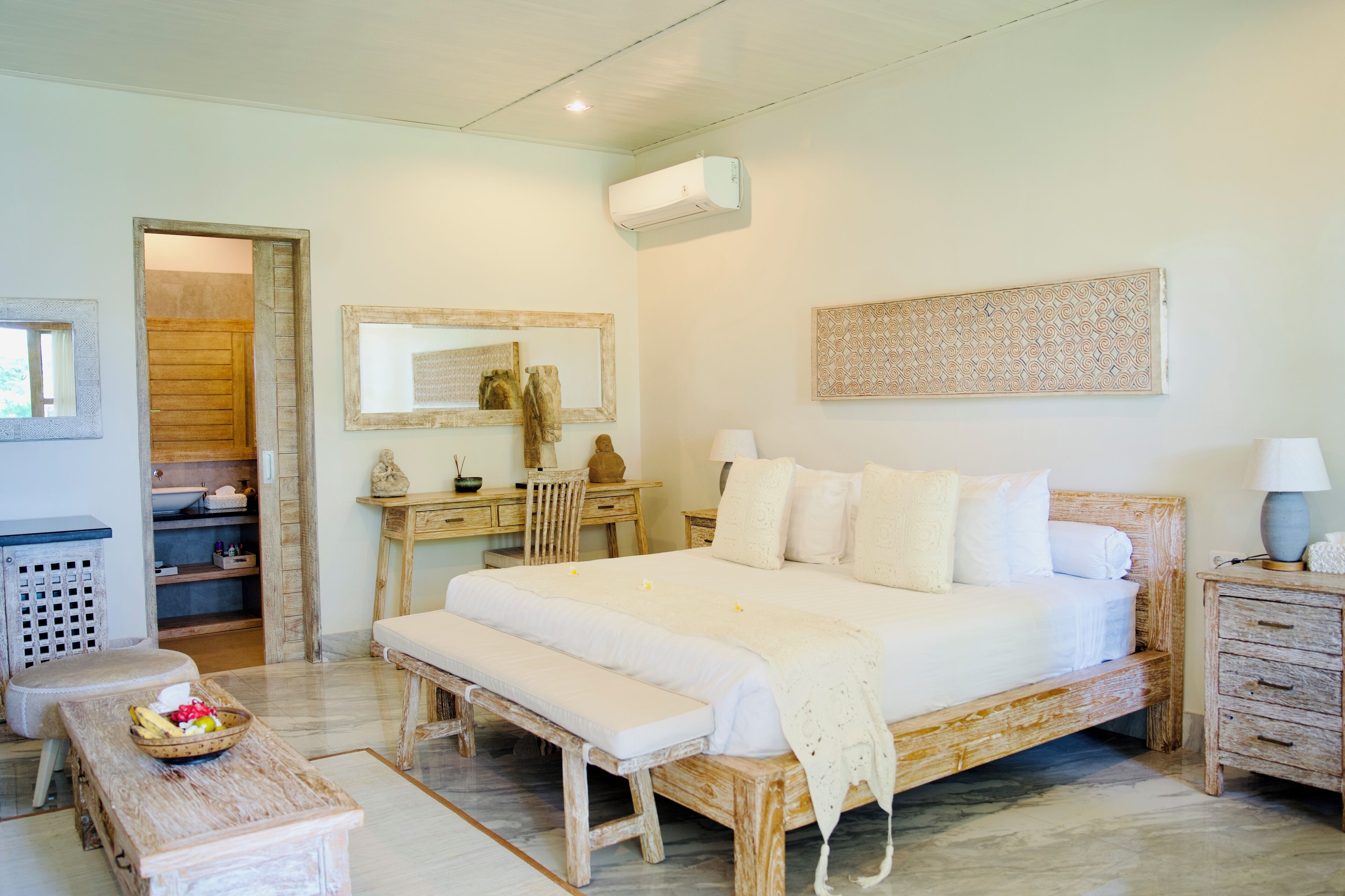A bedroom with a wooden bed, a bedside table, a bench, a desk, a mirror, and an air conditioning unit.
