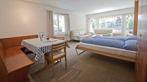 Une pièce de taille modeste comprenant un lit, une petite table avec des chaises, un banc et une fenêtre avec des rideaux.